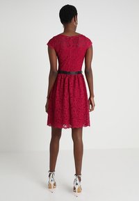 Robe en dentelle rouge avec des manches courtes, un corsage ajusté et une jupe évasée. Elle présente une ceinture noire à la taille et un ourlet à volants.