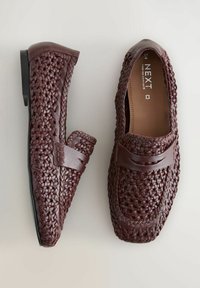 Des mocassins en cuir tressé marron présentent un bout arrondi et une semelle plate noire, avec une bande en cuir sur le dessus pour un détail de design supplémentaire.