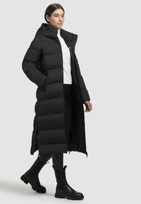 Abrigo de plumas largo negro con capucha, que cuenta con secciones acolchadas y cierre frontal. Combinado con botas de tacón de cuero negro y un suéter blanco de cuello alto.