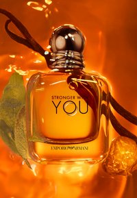 Bouteille de parfum en verre étiquetée "Stronger With You" par Emporio Armani, contenant un liquide ambré, des gousses de vanille et des feuilles dans une lumière orange chaude.
