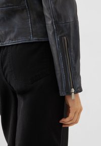 Black leather jacket with a smooth texture, featuring a zippered cuff and tailored fit. Worn with black pants, showcasing stitching details.