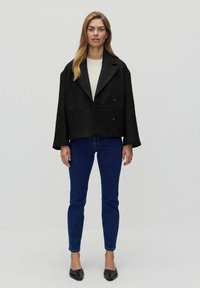 Czarna, oversize'owa, dwurzędowa kurtka z szerokim kołnierzem, z teksturowanej tkaniny, w połączeniu z jasnym swetrem, niebieskimi rurkami i czarnymi balerinami.