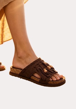 Pied portant une sandale mule en daim marron avec des pompons et des perles en bois sur semelle en liège sur fond uni.