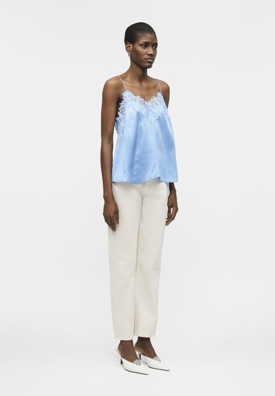 Γυναίκα που φοράει ανοιχτό μπλε σατέν καλοκαιρινό τοπ με δαντέλα, εκρού παντελόνι κατευθείαν και λευκά μυτερά τακούνια με διακοσμητικά, στέκεται πλάγια.