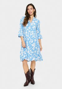 Rochie florală de culoare albastru deschis, cu flori albe, de tip v-neck, cu mâneci clopot până la trei sferturi, fustă evazată, purtată cu cizme de cowboy maro.