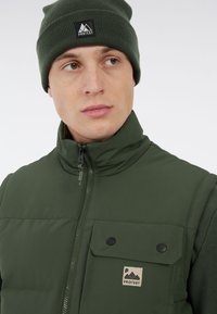 Groene geïsoleerde vest met een hoge kraag, een borstzak met twee knopen en contrasterende stoffen texturen. Gecombineerd met een bijpassende gebreide muts.