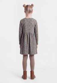 Ragazza giovane vista di spalle, indossa un vestito lungo a maniche lunghe grigio con motivi floreali, collant beige, stivali marroni e i capelli acconciati in due chignon.