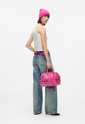 Nő rózsaszín sapkában, fehér ujjú pólóban, bő szárú farmernadrágban áll, rózsaszín táskát tart a kezében, egy sima fehér háttér előtt.