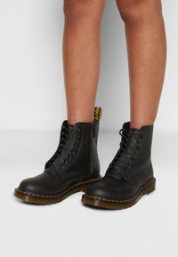 Piernas que llevan botas de tobillo de cuero negro con cremalleras laterales, suelas gruesas, costuras amarillas y tiradores, contra un fondo sencillo.