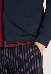 Marine blauwe ritsjack met geribbelde manchetten en zoom, gelaagd over een bordeauxrood shirt, gecombineerd met gestreepte broek in marineblauw, wit en rood.