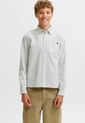 Jeune personne souriante aux cheveux bouclés portant une chemise légère rayée boutonnée et un pantalon beige, debout devant un fond blanc uni.