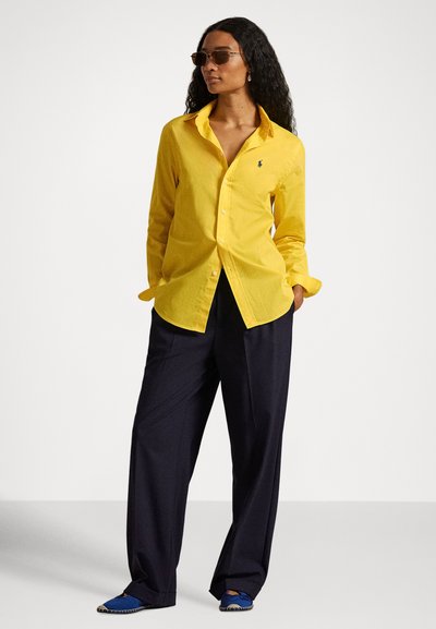 Moteris dėvi geltoną marškinių tipo palaidinę, tamsias laisvas kelnes, mėlynas slip-on stiliaus batus ir akinukus nuo saulės, stovinti su rankomis kišenėse prieš baltą foną.