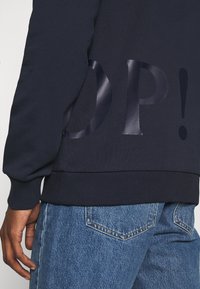 Mörkblå sweatshirt med glänsande "OP!" text på ryggen, bärs med blå denimjeans och visar en vänsterhand och handled.