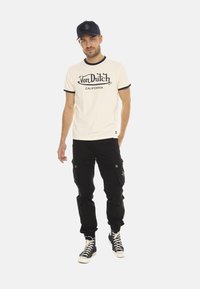 T-shirt blanche avec col noir, portant le texte "Von Dutch California", associée à un pantalon cargo noir et des baskets montantes. Tenue décontractée.