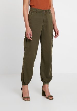 Personne portant un pantalon cargo vert olive avec des poignets élastiques et des sandales à talons assorties, associé à un haut couleur rouille.