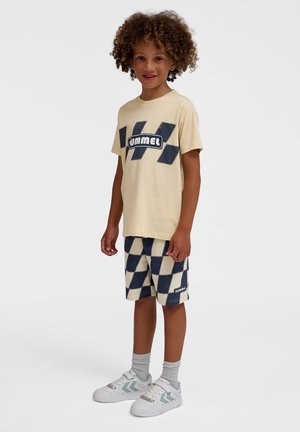 Lys gul t-shirt med et blåt logo, parret med ternede beige og marineblå shorts. Hvide sneakers med blå og grå detaljer. Krøllet hår.