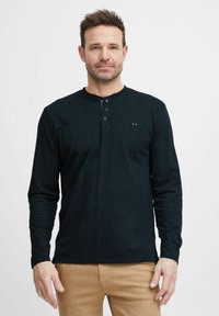 Pikkade varrukatega must henley-särk, millel on kolme nupuga eesmine tõmblukk, pehme tekstuur ja väike logo detail rindkere piirkonnas, kombineeritud heledate chinos-pükstega.