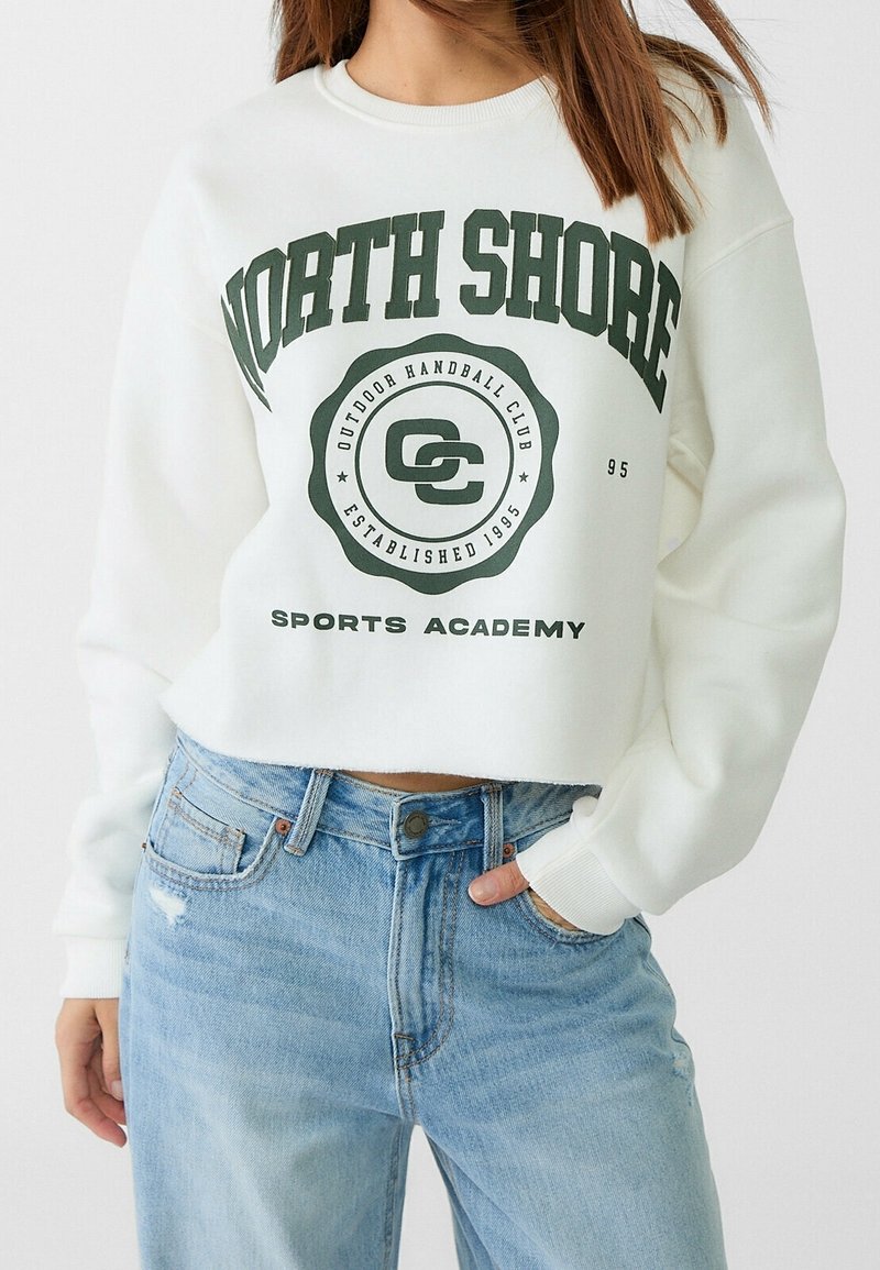 Femme portant un sweat-shirt court blanc avec le texte "North Shore Outdoor Handball Club Established 1995 Sports Academy" et un jean bleu clair.