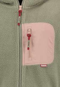 Chaqueta de forro polar verde con un bolsillo en el pecho con cremallera rosa y tiradores de cremallera a juego con cordones rojos.