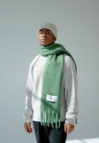 Groene wollen sjaal met franjes aan de randen, merklabel, gedrapeerd over een lichtkleurige trui, gedragen met een beige gebreide muts.