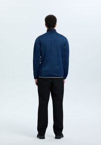 Giacca mezze zip texture blu navy con colletto alto, abbinata a pantaloni neri e scarpe nere, vista da dietro.