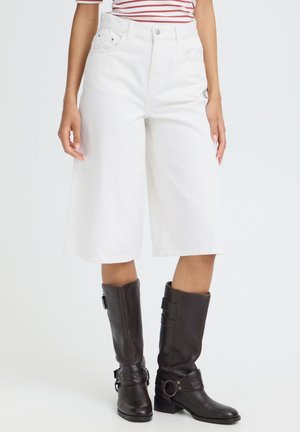 Personne portant un short blanc ample mi-cuisse, un haut rayé et des bottes en cuir marron foncé montant jusqu'aux genoux, debout devant un fond uni.