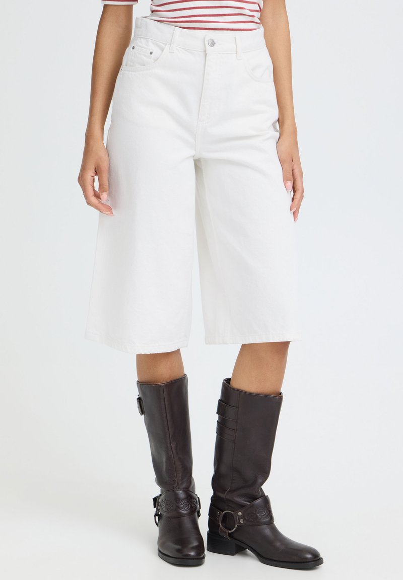 Personne portant un short blanc ample mi-cuisse, un haut rayé et des bottes en cuir marron foncé montant jusqu'aux genoux, debout devant un fond uni.