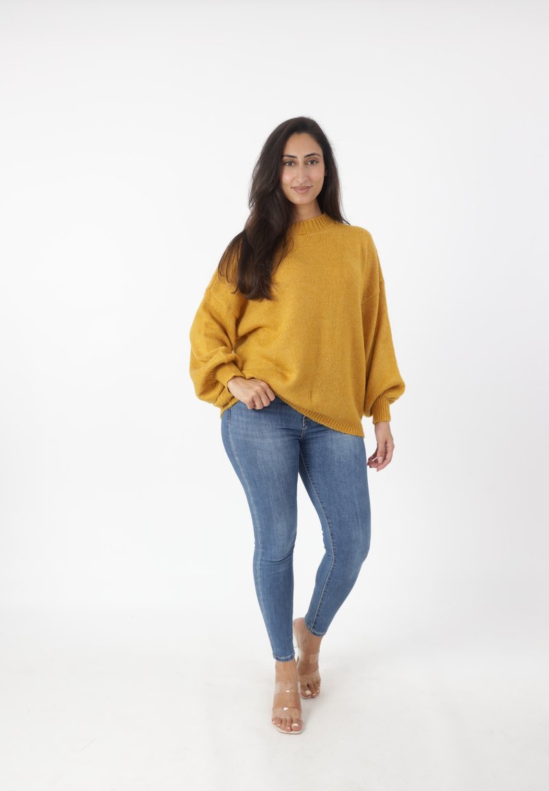 Musztardowy, oversize'owy sweter dzianinowy z wysokim kołnierzem, zestawiony z jasnobłękitnymi rurkami i przezroczystymi sandałami na obcasie, stojący na tle białym.