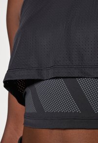 Czarny krótki top sportowy z perforowaną teksturą, nałożony na dopasowaną bazową warstwę z geometrycznym wzorem w kropki w odcieniach szarości.