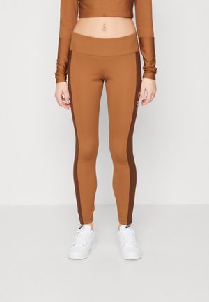 Zbliżenie osoby noszącej brązowe legginsy z ciemniejszymi paskami po bokach i białe buty sportowe Nike, stojącej na jednolitym tle.