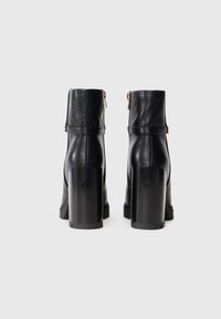 Botines de tacón negro con un cuerpo de cuero suave, puño de cuero con estampado, detalle de cremallera dorada y tacones altos y voluminosos.