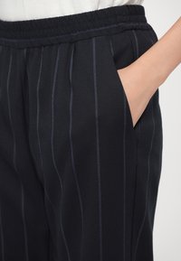 Main dans la poche d'un pantalon noir à fines rayures verticales et taille élastique, porté avec un haut blanc.