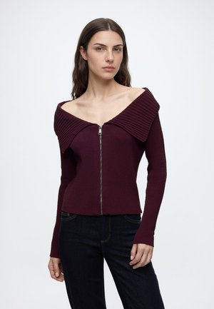 Frau mit langen dunklen Haaren trägt einen dunkelvioletten schulterfreien Reißverschluss-Pullover und dunkle Jeans und steht vor einem schlichten weißen Hintergrund.