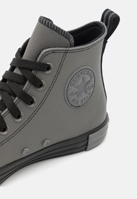 Sneaker alto grigio in tela testurizzata. Presenta lacci neri, suola in gomma e una toppa circolare con logo sul lato.