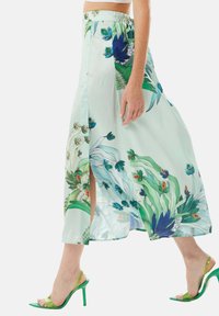 Long, light green floral skirt featuring blue and orange botanical patterns, with a side slit and elastic waistband. Paired with green heels.