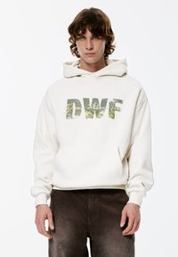 Crèmekleurige hoodie met een kangoeroezak, met de letters "DWF" in een getextureerde groene print met gouden accenten, gedragen met bruine corduroy broek.