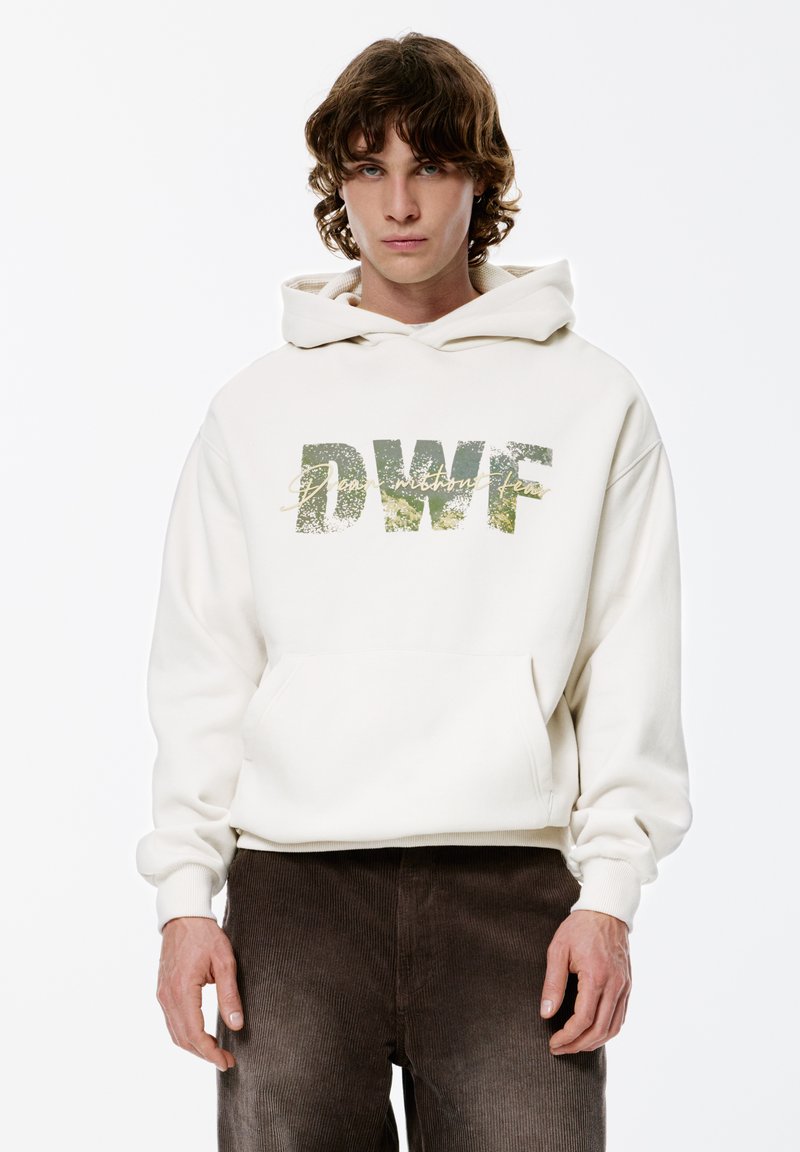 Crèmekleurige hoodie met een kangoeroezak, met de letters "DWF" in een getextureerde groene print met gouden accenten, gedragen met bruine corduroy broek.