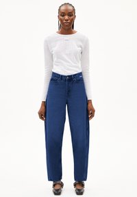 Donkerblauwe high-waisted jeans met een rechte pasvorm, gecombineerd met een witte top met lange mouwen en een subtiele merklabel. Glad textuur.