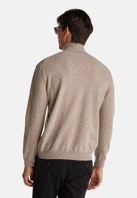 Beige Pullover mit hohem Kragen und geripptem Saum. Weicher Stoff, lange Ärmel und schlanker Schnitt. Getragen mit schwarzen Hosen.