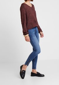 Top de manga larga con estampado animal rojo y escote en V, combinado con jeans ajustados de color azul claro con detalle de cremallera en el tobillo y mocasines negros.