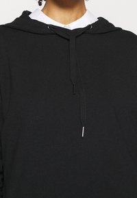 Haut à capuche en coton noir avec un cordon de serrage, présentant des embouts de couleur argentée. Texture douce, design décontracté et sans motifs visibles.