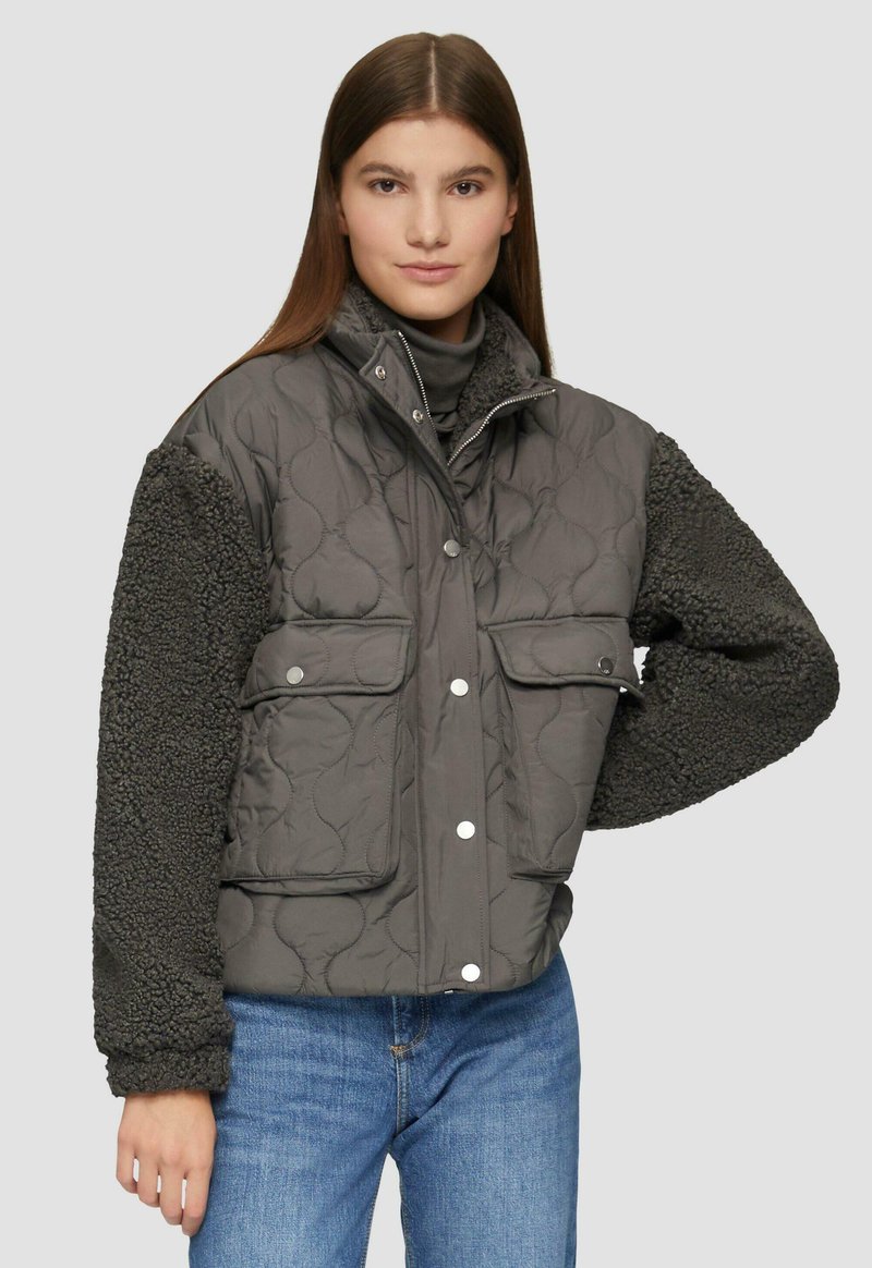 Vrouw in een grijs gewatteerd jasje met gestructureerde mouwen en voorzakken, over blauwe jeans, staande tegen een simpele lichte achtergrond.