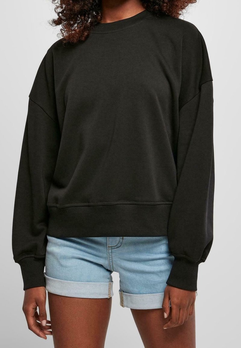 Femme portant un sweat-shirt noir à manches longues et un short en denim bleu clair retroussé sur un fond uni.