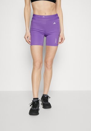 Frau trägt lilafarbene Adidas-Sportshorts und schwarze Sneakers, steht vor einem schlichten hellen Hintergrund.