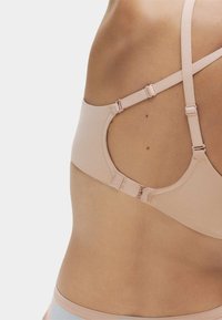 Rückansicht einer Person, die einen beigen BH mit verstellbaren Trägern und Hakenverschluss vor einem schlichten hellen Hintergrund trägt.