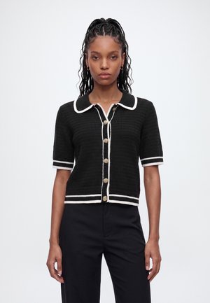 Femme aux cheveux tressés portant un cardigan noir à manches courtes boutonné avec une bordure blanche, associé à un pantalon noir, debout devant un fond blanc uni.
