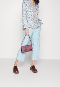 Blau gemusterte Bluse mit Rüscheärmeln, hellblaue Schlaghose und bordeauxfarbene Lederclogs. Hält eine kleine bordeauxfarbene Handtasche.