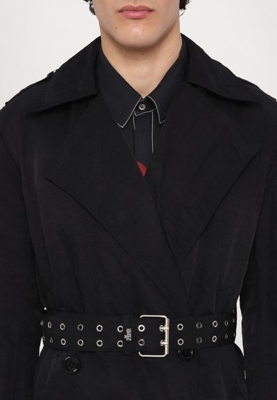 Schwarzer Trenchcoat mit strukturiertem Finish, breiten Revers, doppelreihigen Knöpfen und einem schwarzen Gürtel mit silbernen Ösen und Schnalle.