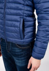 Giacca puffer navy con design trapuntato, chiusura frontale con zip e tasche laterali. Indossata sopra jeans in denim, con polsini a coste.