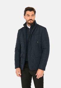 Giacca trapuntata blu navy con un colletto alto, multiple tasche e chiusura con zip. Realizzata in un tessuto morbido e testurizzato con una vestibilità su misura.